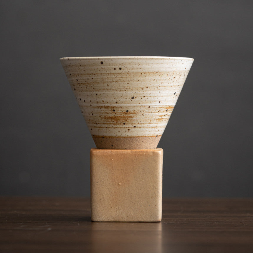 Beigefarbene Keramiktasse mit horizontalem Linienmuster und feinen Sprenkeln. Die konische Tasse steht auf einem eckigen Sockel aus naturbelassener Grobkeramik. Handgefertigtes Design im japanisch minimalistischen Stil.