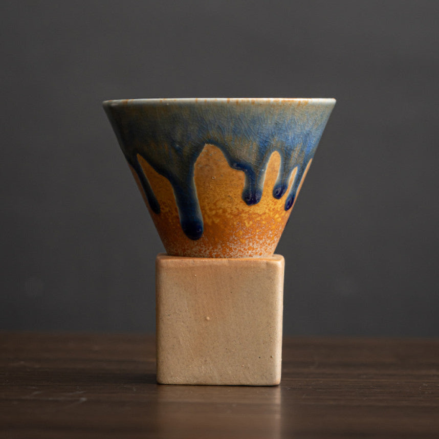 Konische Keramiktasse mit leuchtend blauer Laufglasur auf orangebraunem Grund. Die handgefertigte Tasse steht auf einem eckigen Sockel aus naturfarbenem Ton. Einzigartiges japanisches Design für moderne Kaffeeästhetik.