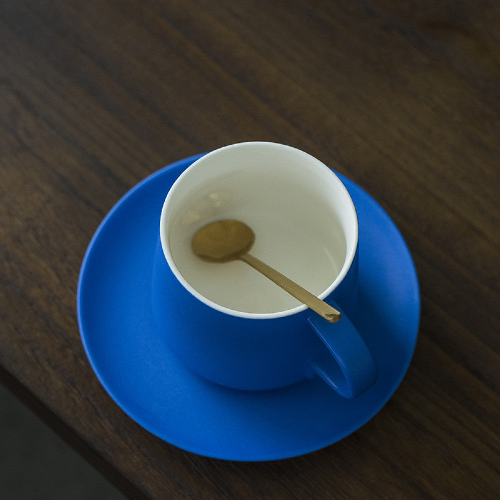 Blaue Porzellantasse mit passendem Unterteller und goldenem Löffel auf dunklem Holz – perfekt für puristische Kaffeeästheten.