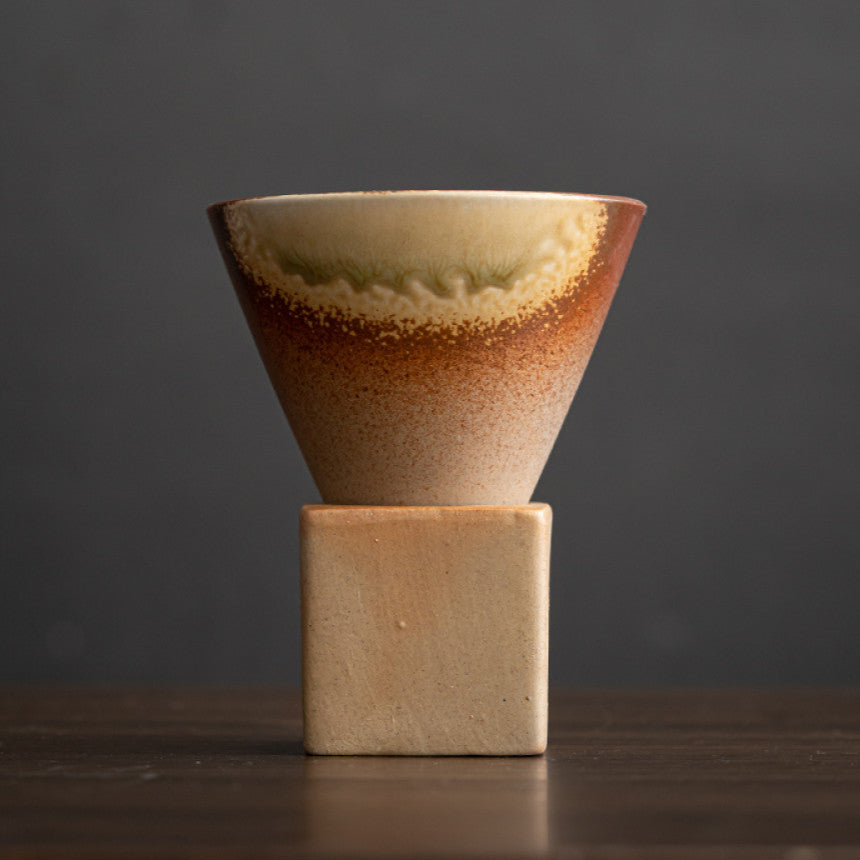Keramiktasse mit cremefarbener Glasur und braunem Verlauf auf konischem Korpus. Feine Wellenstruktur unter dem Rand verleiht der Tasse eine besondere Textur. Der kubische Sockel aus naturbelassenem Ton rundet das japanisch inspirierte Design ab.