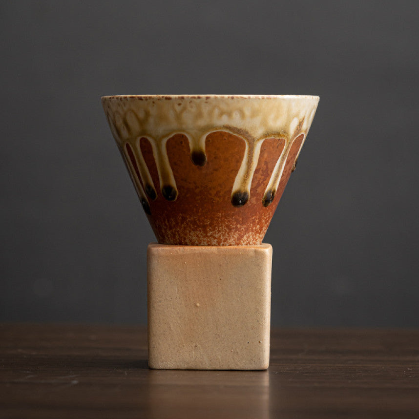Braune Keramiktasse mit heller Laufglasur und dunklen Akzenten. Die konische Form sitzt auf einem eckigen Sockel aus naturbelassenem Ton. Charaktervolles Design im japanischen Stil für Espresso oder Latte.