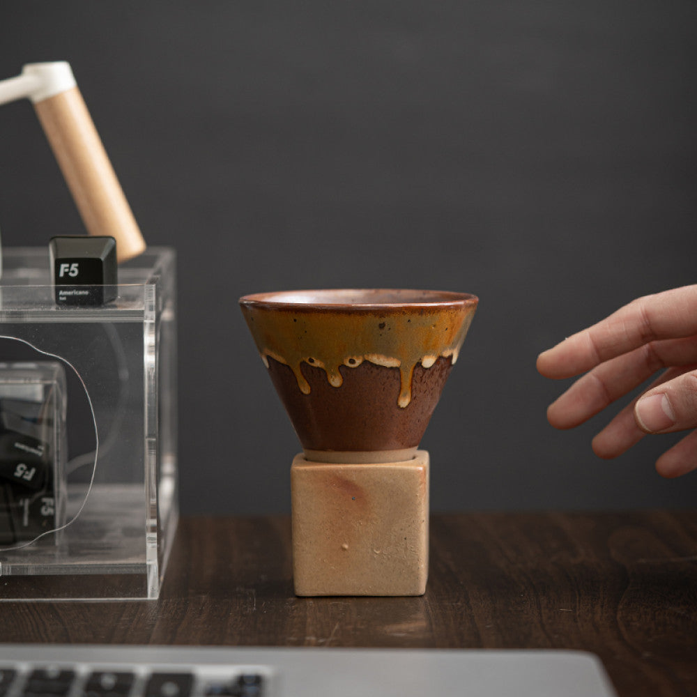 Braune Keramiktasse mit gelblicher Glasur auf kubischem Sockel aus Naturton auf einem Schreibtisch. Eine Hand greift nach der Tasse neben einem Keyboard und Designobjekt. Stilvolle Kombination aus Handwerk und modernem Workspace.