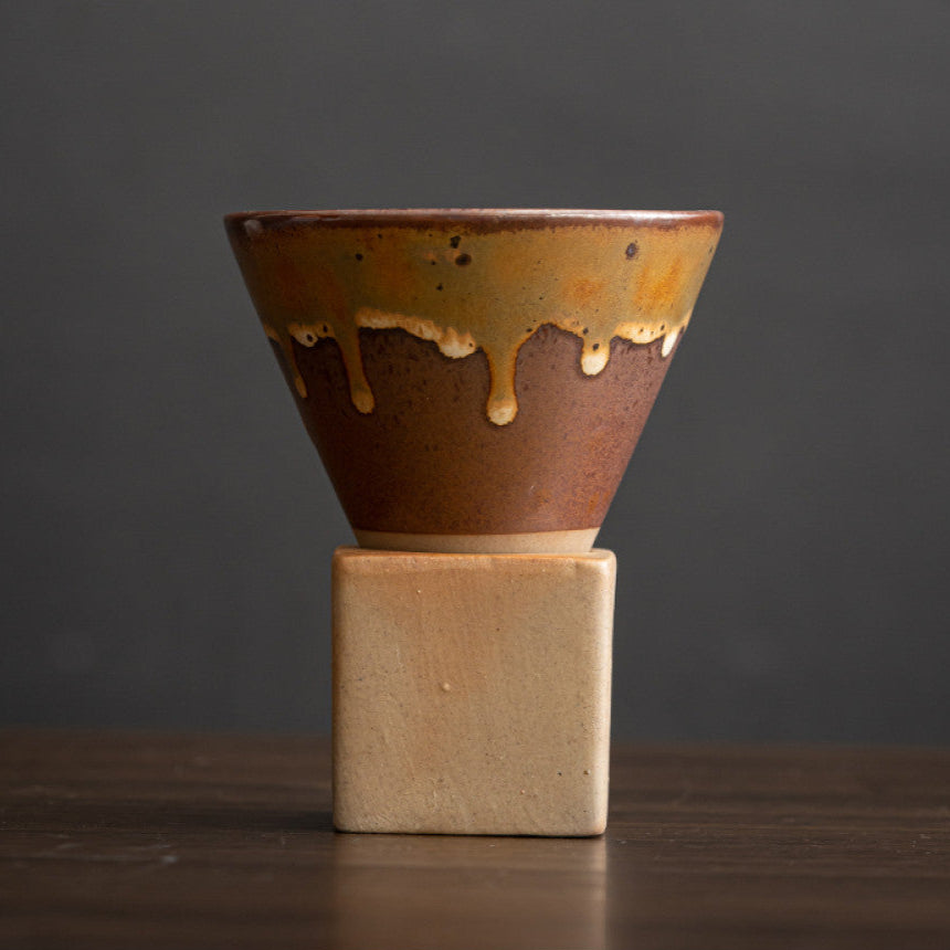 Keramiktasse mit brauner Oberfläche und gelblicher Laufglasur im handgefertigten Look. Die konische Form ruht auf einem eckigen Sockel aus naturfarbenem Ton. Ausdrucksstarkes Design im japanischen Retro Stil.