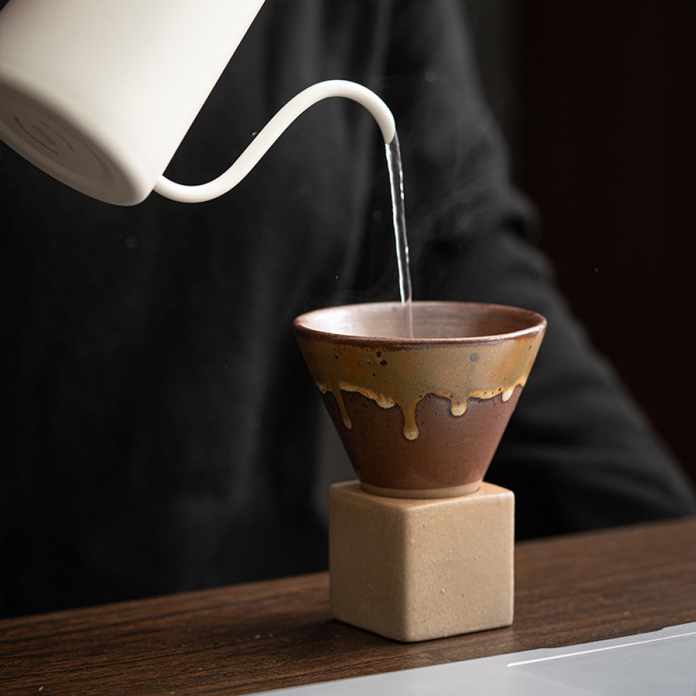Heisses Wasser wird aus einem weissen Kessel in eine braun glasierte Keramiktasse mit konischer Form gegossen. Die Tasse steht auf einem eckigen Sockel aus Naturton und eignet sich ideal für Pour-Over Kaffee oder Tee.