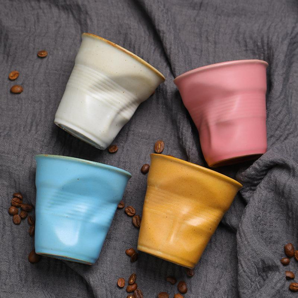 Keramiktassen in Rosa, Weiss, Schwarz, Gelb und Blau auf einem Leinentuch mit verstreuten Kaffeebohnen. Stylisches Arrangement mit kreativer Farbwahl.