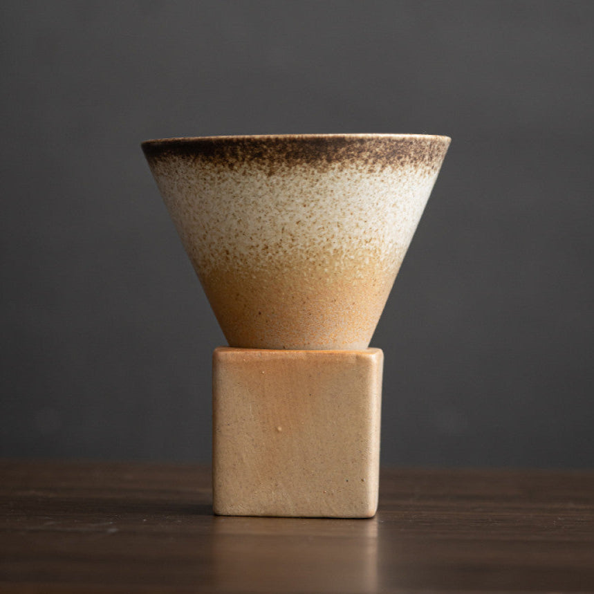Helle Keramiktasse mit braunem Farbverlauf auf naturbelassenem Ton. Die konische Form steht auf einem eckigen Sockel aus Grobkeramik. Schlichtes japanisches Design für stilvollen Kaffeegenuss.