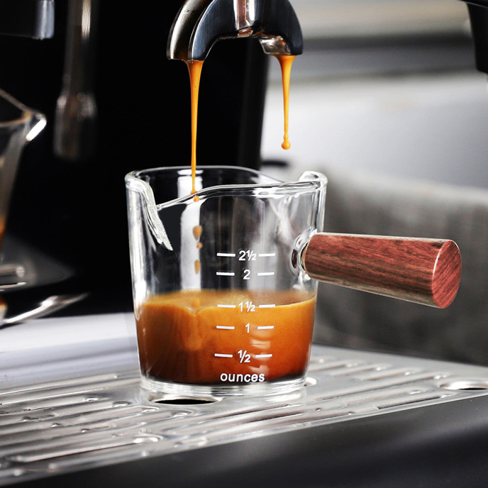 Doppelter Espresso fliesst in ein klares Espressoglas mit Holzgriff unter einer Siebträgermaschine. Skala in Unzen für präzises Abmessen. Perfekt für Barista Shots und Kaffeegenuss mit Stil.