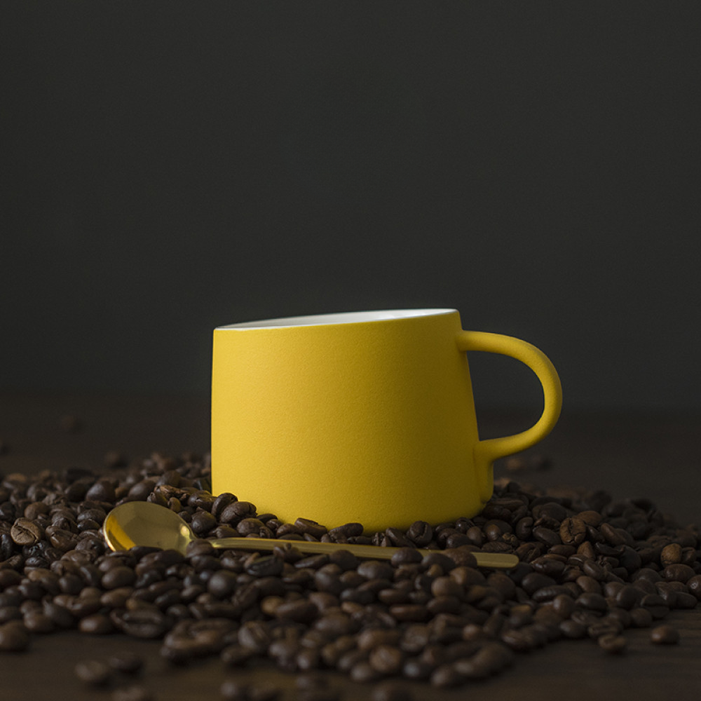 Gelbe Porzellantasse mit passendem Unterteller auf frisch gerösteten Kaffeebohnen – stylische Präsentation mit Charakter.
