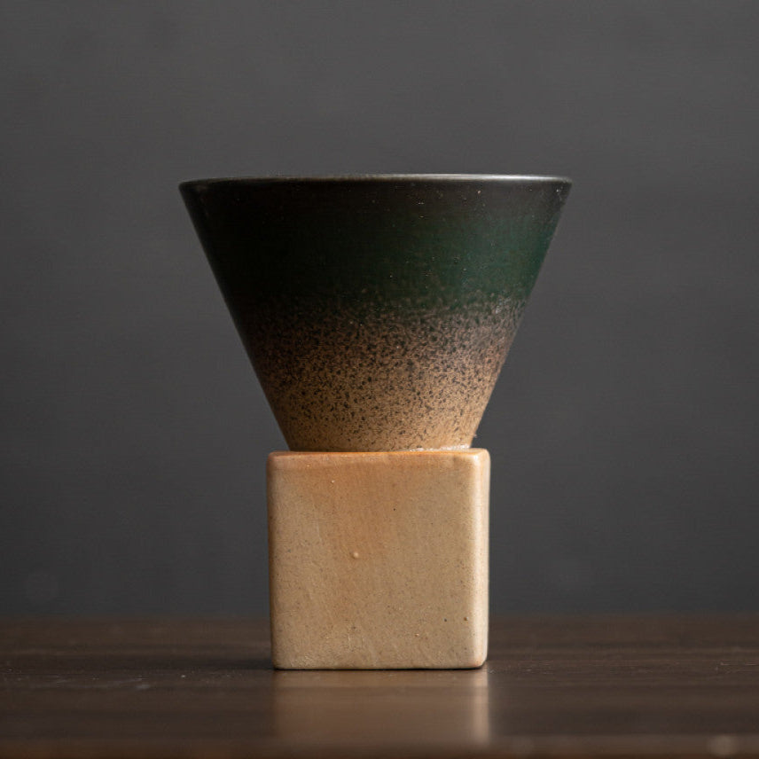 Dunkelgrüne Keramiktasse mit feinem Farbverlauf zu naturbelassenem Ton. Die konische Tassenform ruht auf einem eckigen Sockel aus heller Grobkeramik. Modernes Design im japanisch minimalistischen Stil.