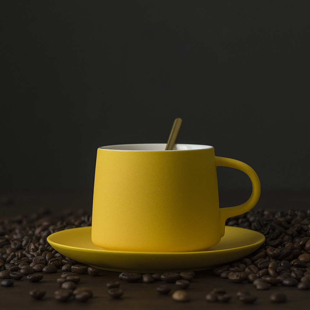 Moderne gelbe Porzellantasse mit goldenem Löffel auf Kaffeebohnen – trendiges Accessoire für Designliebhaber.