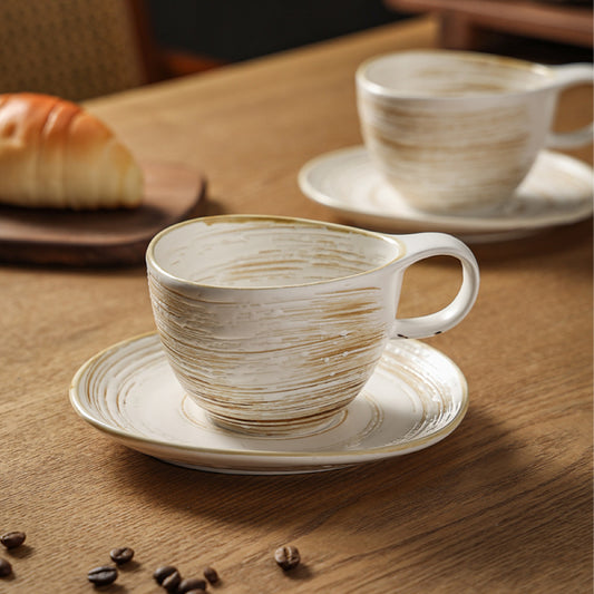 Set aus zwei beigen Keramiktassen mit Untertassen auf Holztisch – perfektes Duo für einen gemütlichen Frühstücksmoment.