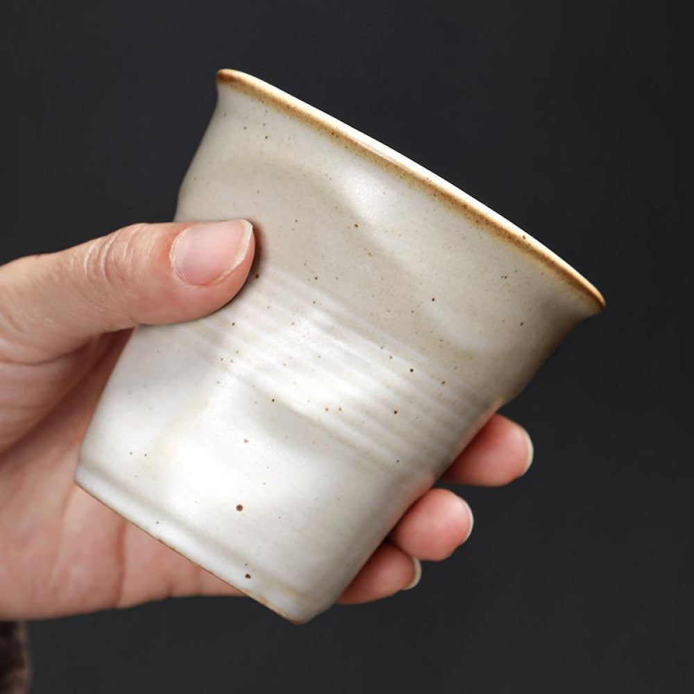 Eine weisse Tasse mit Knickoptik wird in der Hand gehalten. Das matte Finish und die organische Form unterstreichen das handgemachte Design.