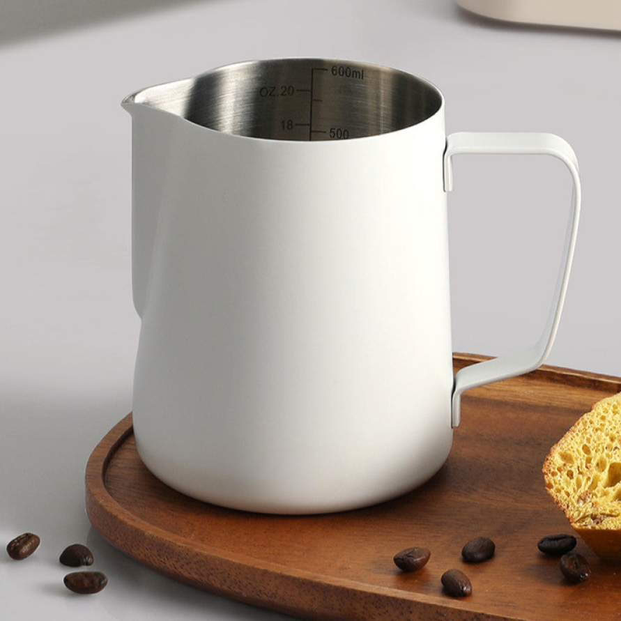 Weisses Milchkännchen aus Edelstahl mit klarer Messskala, platziert auf einem Holztablett mit Kaffeebohnen. Elegantes Zubehör für Barista-Momente.