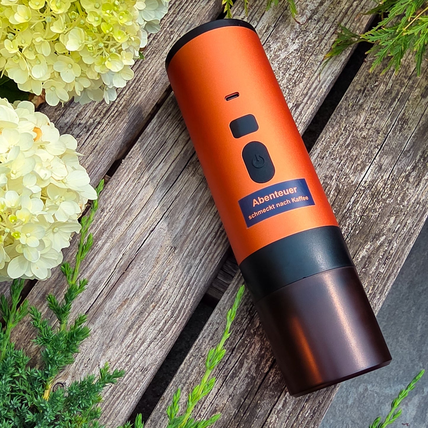 Die orange Honeybean Kaffeemaschine liegt auf einer Holzbank zwischen weissen Blüten und grünen Pflanzen – ideal für deinen Outdoor-Kaffeegenuss.