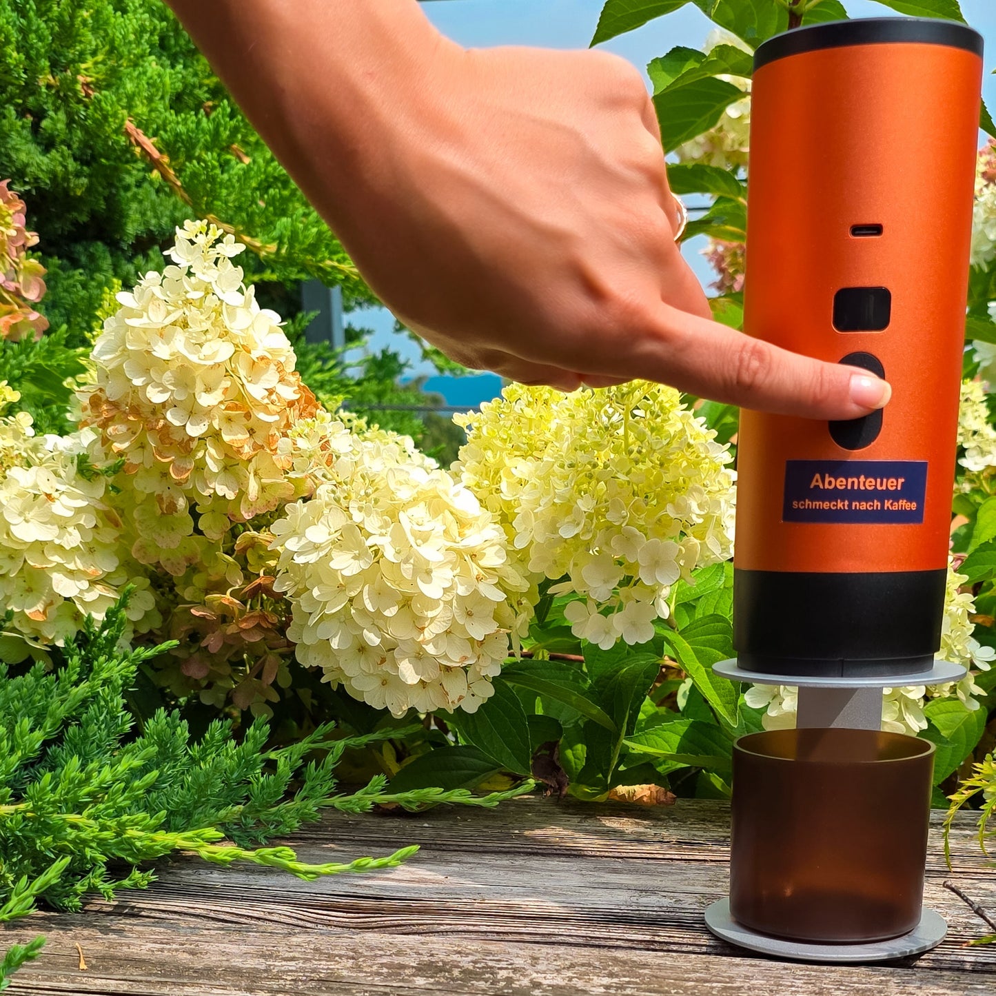 Eine Hand drückt den Einschaltknopf einer orangen Honeybean Kaffeemaschine auf einer Holzbank inmitten von weissen Hortensien und grünem Garten.