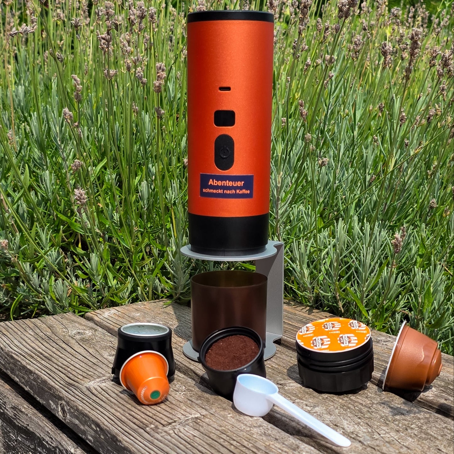Outdoor-Aufnahme einer orangen kabellosen Kaffeemaschine von Honeybean mit Kaffeekapseln, Löffel und Zubehör auf einer Holzbank im Garten vor Lavendel.