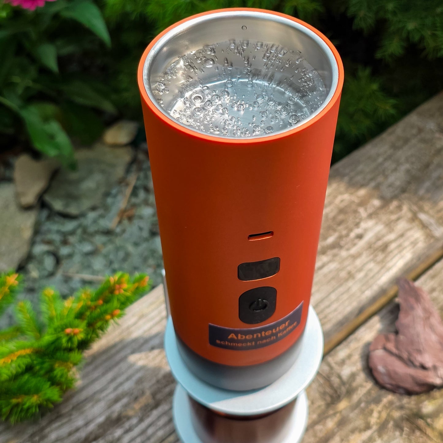 Nahaufnahme der geöffneten orangen Honeybean Kaffeemaschine mit sprudelndem Wasser beim Aufheizen, platziert auf einer Holzbank im Garten.