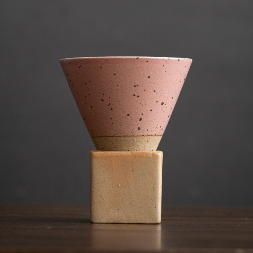 Minimalistische Keramiktasse in Rosa mit schwarzer Sprenkeloptik und naturfarbenem Fuss. Die konische Form sitzt auf einem eckigen Sockel aus Grobkeramik. Handgefertigtes Design im japanischen Stil für stilvollen Kaffeegenuss.