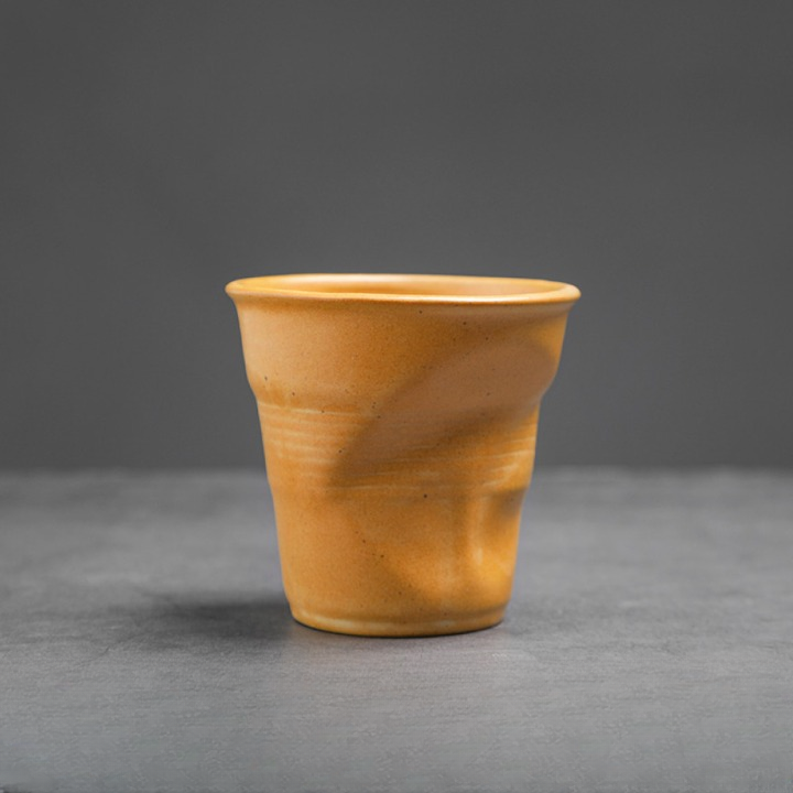 Gelbe Keramiktasse mit organischem Knickdetail auf grauem Untergrund. Schlichtes Design mit Charakter.