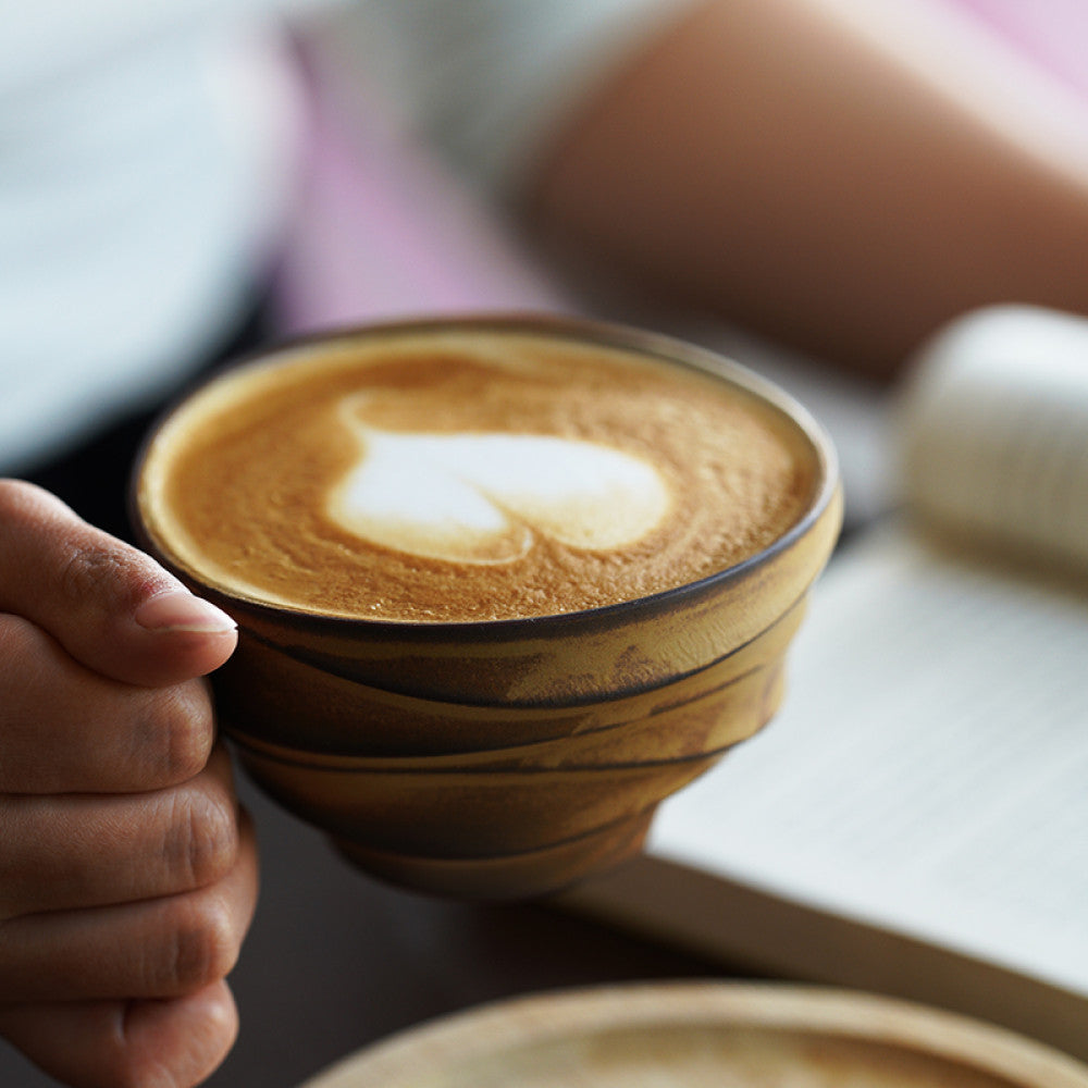 Detailaufnahme einer handgehaltenen Keramiktasse mit geschwungenem Henkel und Latte Art.