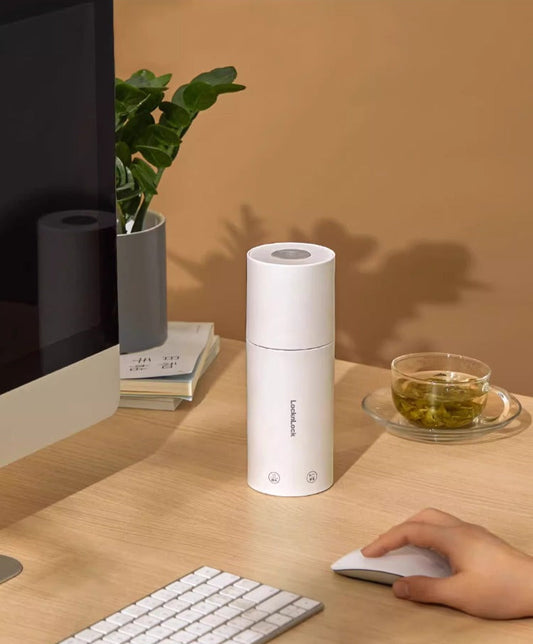 Weisser akkubetriebener Wasserkocher auf einem Holzschreibtisch neben einer Tasse Tee, einer Computertastatur, einer Maus und einem Monitor. Im Hintergrund steht eine Pflanze auf einem Stapel Bücher.