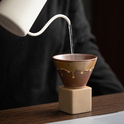 Heisses Wasser wird aus einem weissen Kessel in eine braun glasierte Keramiktasse mit konischer Form gegossen. Die Tasse steht auf einem eckigen Sockel aus Naturton und eignet sich ideal für Pour-Over Kaffee oder Tee.