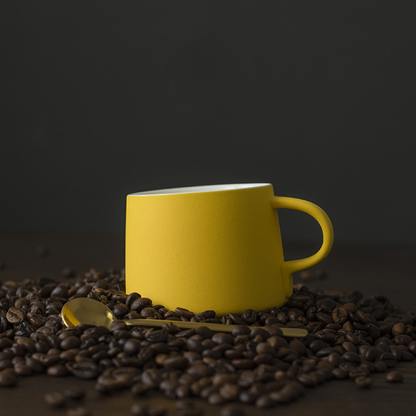 Gelbe Porzellantasse mit passendem Unterteller auf frisch gerösteten Kaffeebohnen – stylische Präsentation mit Charakter.