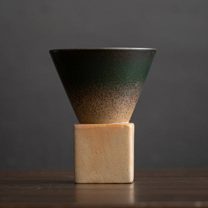 Dunkelgrüne Keramiktasse mit feinem Farbverlauf zu naturbelassenem Ton. Die konische Tassenform ruht auf einem eckigen Sockel aus heller Grobkeramik. Modernes Design im japanisch minimalistischen Stil.