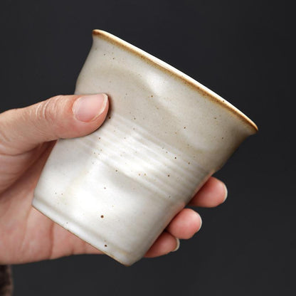 Eine weisse Tasse mit Knickoptik wird in der Hand gehalten. Das matte Finish und die organische Form unterstreichen das handgemachte Design.