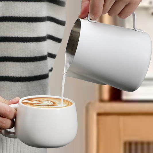 Milchschaum wird mit einem weissen Edelstahlkännchen in eine Tasse Cappuccino gegossen. Perfekte Anwendung für Latte Art Zuhause oder im Café.