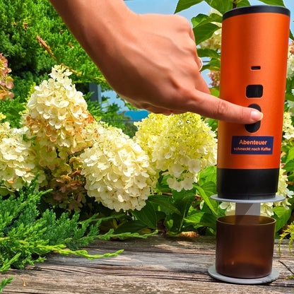Eine Hand drückt den Einschaltknopf einer orangen Honeybean Kaffeemaschine auf einer Holzbank inmitten von weissen Hortensien und grünem Garten.