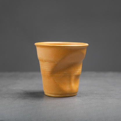 Gelbe Keramiktasse mit organischem Knickdetail auf grauem Untergrund. Schlichtes Design mit Charakter.