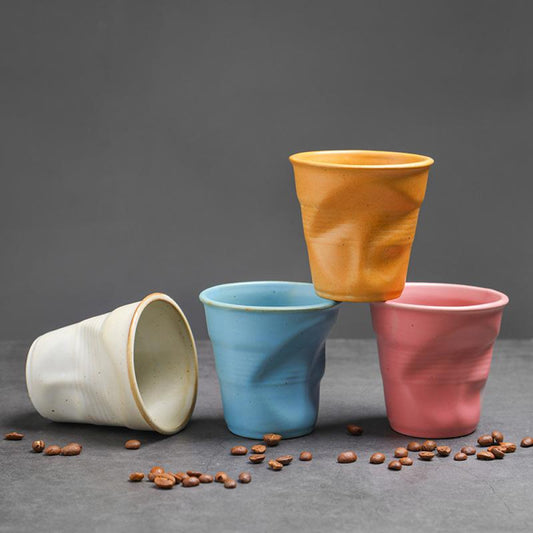 Vier Keramiktassen mit Knickoptik in Blau, Rosa, Gelb und Weiss liegen dekorativ mit Kaffeebohnen auf grauem Untergrund. Modernes Design in trendigen Farben.