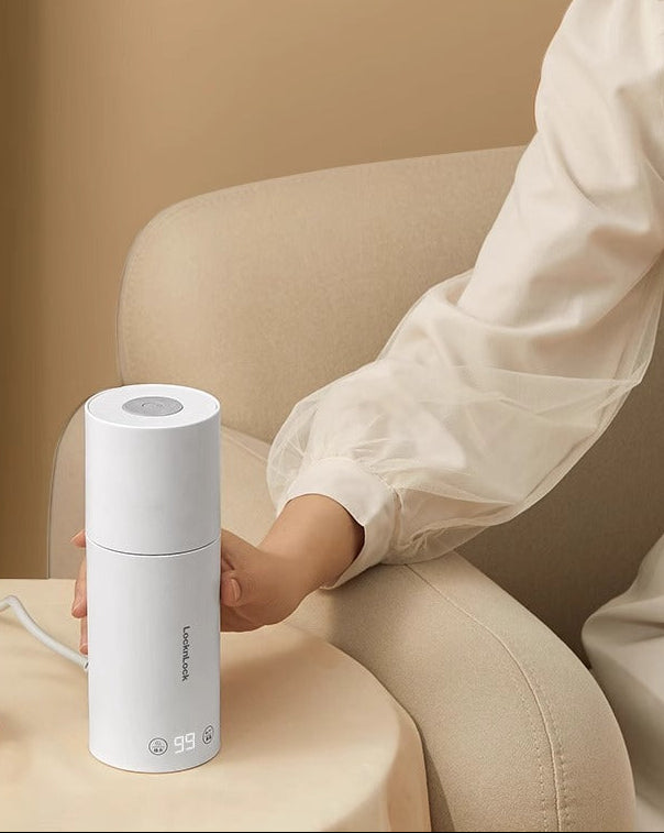 Eine Person berührt einen weissen Wasserkocher mit digitalem Display, der auf einem runden Tisch neben einem beigen Polstersessel steht. Das Display zeigt 99 Grad an.